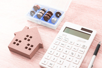 A wooden house cutout, a white calculator, a pen, and a container of coins are arranged on a textured surface, suggesting financial planning for a home.