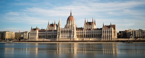 The image features a beautiful scene of a large city with multiple tall buildings, including a majestic castle in Budapest, Hungary. In front of the castle, there is a river. Boats can be seen on the water, adding to the picturesque atmosphere of the city. The scene showcases the charm and grandeur of the city, making it an attractive destination for tourists and locals alike.