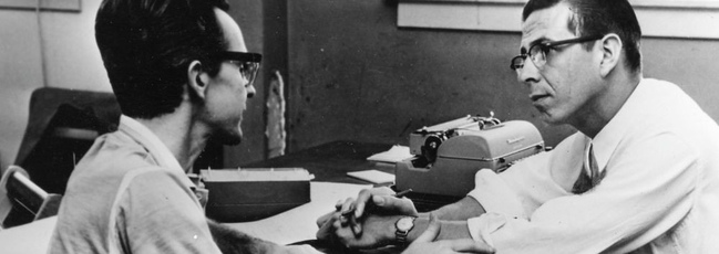 A man is using a typewriter while a businessman watches him.
