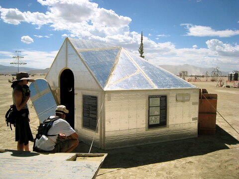 A group of people are standing outside a house that appears to be made of solar panels. The house seems to be a unique structure, possibly designed for energy efficiency or to showcase solar technology. The group consists of four individuals, with a couple of backpacks and a handbag among them, possibly carrying their belongings for a trip or event. They seem to be standing close to the house, examining it and possibly admiring its innovative design.