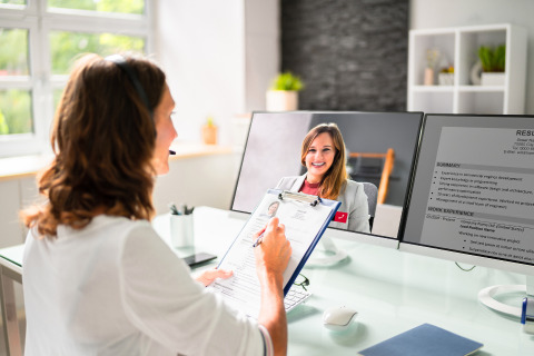 A person with a headset conducts a video interview, looking at a resume on a clipboard and a screen displaying the candidate's face.