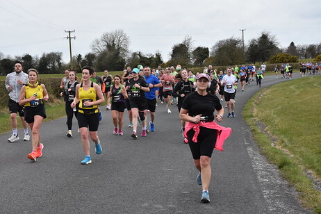 A woman is running down a road, holding a pink water bottle. There are many other people in the scene, running alongside her.