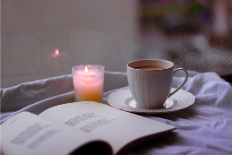 An open book rests on a textured surface next to a steaming cup of tea on a saucer and a lit candle.