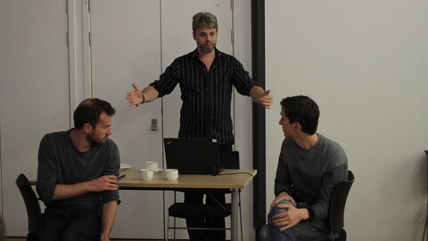 Three men are sitting at a table in a room. One man is standing in the room and pointing at something. The other two men are sitting at the table, engaged in a discussion. A laptop is on the table between the two sitting men. A cup is also placed on the table.