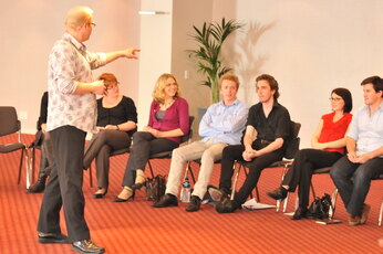 An older gentleman is giving a presentation to a group of people, who are sitting in chairs arranged in a semi-circle. The chairs are positioned in various directions, with some facing the presenter and others facing in the opposite direction. A potted plant is located near the center of the room, and a bottle can be seen on the floor, closer to one of the chairs. In addition to the chairs, there are also a couple of handbags and a cell phone in the scene.