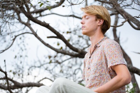 A man with brown hair is sitting under a tree, looking deep in thought