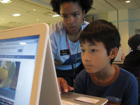 A young boy looking at his laptop while a woman watches and teaches him.