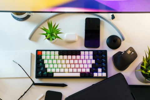 A top-down view of a desk setup with a mechanical keyboard, smartphone, wireless mouse, tablet, and small potted plants.