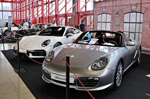 A large grey car is parked in a room with other cars around it. It is in front of a brick wall.