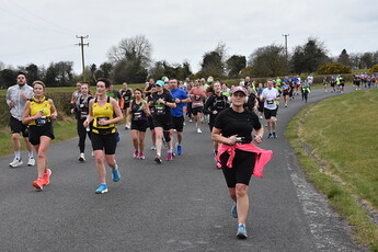 A woman is running down a road, holding a pink water bottle. There are many other people in the scene, running alongside her.