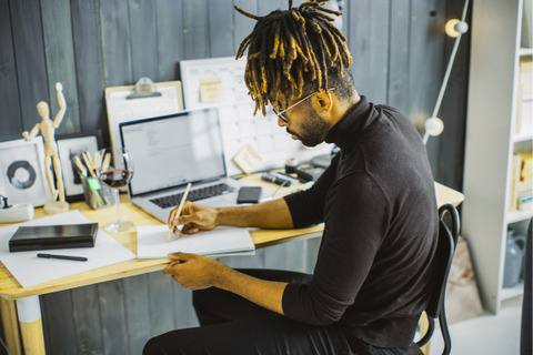 A person sits at a desk with a laptop, glass of wine, and art supplies, writing in a notebook.
