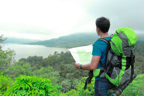 A person wearing a large green backpack looks at a map overlooking a lake and forested hills under a cloudy sky.