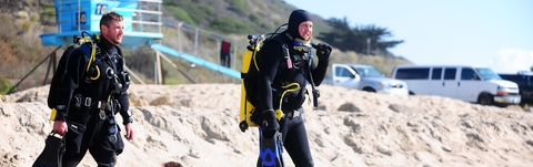 Two men are standing on the beach with scuba gear.