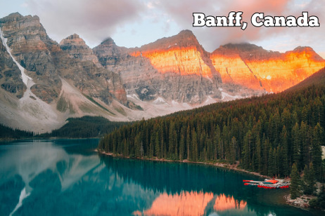 Sunrise illuminates the peaks of a mountain range overlooking a dense pine forest and a turquoise lake with a cluster of red canoes docked at the shore.