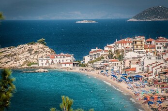 A small island with a few houses and a large rock next to a sandy beach filled with blue umbrellas and chairs.