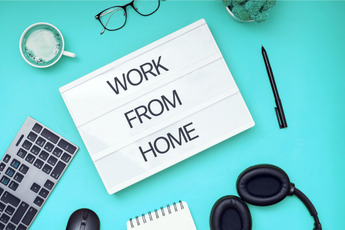 Overhead shot of a desk setup with a keyboard, mouse, notebook, headphones, pen, glasses, mug, and a sign that reads "WORK FROM HOME" on a teal background.