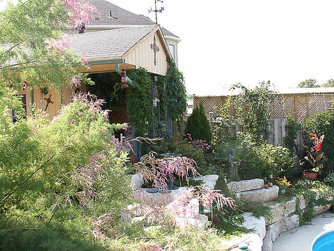 A view of a beautiful garden with pink flowers and green trees. The garden also includes a pond in the middle and a house nearby.