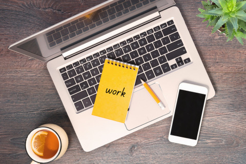 A laptop, smartphone, notepad, pencil, cup of tea, and a small potted plant arranged on a wooden desk.