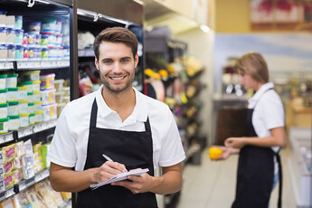 A man is standing behind a deli counter, smiling, with a clipboard in hand.