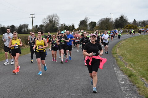 A woman is running down a road, holding a pink water bottle. There are many other people in the scene, running alongside her.