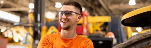A man wearing a yellow shirt and goggles is standing in a factory.