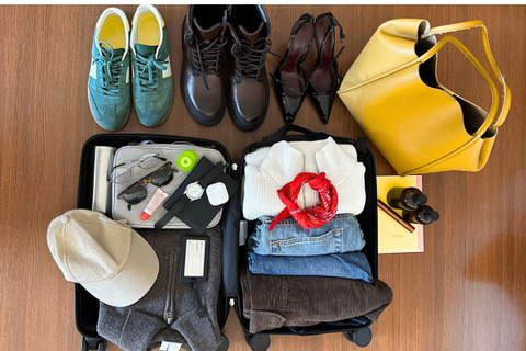 Overhead shot of an open suitcase packed with folded clothes, toiletries, and shoes, next to a yellow bag and a pair of boots, all on a wooden floor.