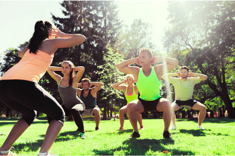 A group is exercising in a park, performing squats with hands behind their heads, led by an instructor.