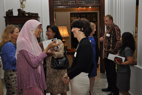 A group of people are gathered in a room, engaged in conversation with one another. The group is diverse, consisting of both men and women, some wearing ties, and one man is holding a cell phone. The conversation appears to be casual and friendly, as they stand close to each other. The room appears to be a hotel lobby or a conference space, with several items present, such as a potted plant, a clock, and a book. Some of the people in the group are holding small items like handbags and a cell phone.