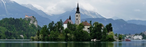 The scene shows a small island in the middle of a lake. On the island, there is a white castle surrounded by trees, adding to the beauty of the area. The serene environment is further accentuated by the presence of a few boats scattered around the lake, giving the impression of a peaceful retreat.