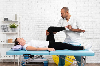 A man in a white shirt and black pants is tending to a woman lying on a table. The man is adjusting the woman's knee and ankle using a knee brace, providing professional care for her health.