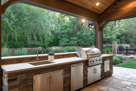 An outdoor kitchen with a stone base and stainless steel appliances sits under a wooden pergola overlooking a lush green backyard.