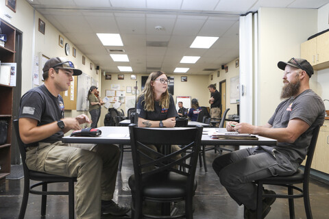 Four people are gathered around a table in an office setting, engaging in conversation. One man is sitting on the left side of the table, while another sits on the right side. Two more people are sitting on the opposite side of the table, engaged in discussion. The table is surrounded by multiple chairs, with one chair visible near the left side of the table and two more chairs on the right side. The scene depicts a casual and social workplace setting with employees interacting and collaborating with one another.