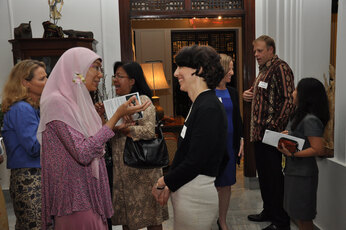 A group of people are gathered in a room, engaged in conversation with one another. The group is diverse, consisting of both men and women, some wearing ties, and one man is holding a cell phone. The conversation appears to be casual and friendly, as they stand close to each other. The room appears to be a hotel lobby or a conference space, with several items present, such as a potted plant, a clock, and a book. Some of the people in the group are holding small items like handbags and a cell phone.