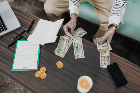 A person is counting money on a wooden table with a laptop, notebook, pens, coins, a lit candle, and a smartphone nearby.