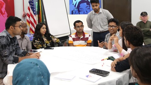 A group of people gathered around a table, working on a project or presentation. There are a few cellphones, a keyboard, a mouse, and a notebook on the table. The people appear to be of Indian descent and are engaged in discussion or collaborating on a task.