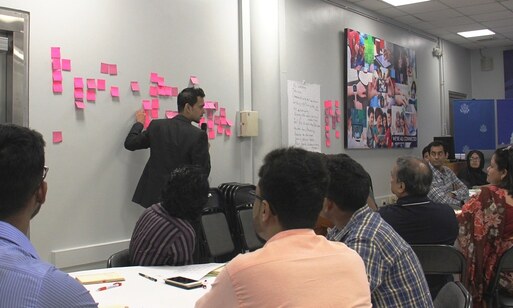 A man teaching a group of people in a conference room, with many notes posted on a wall.